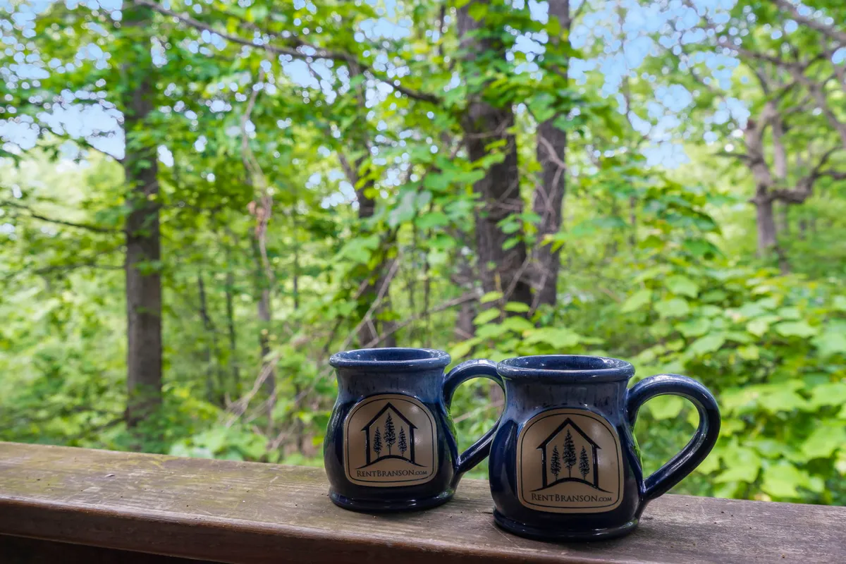 The photo features two coffee mugs perched on a wooden railing overlooking thick green foliage. The scene suggests a tranquil moment spent outdoors, immersed in nature, and hints at relaxing with a coffee while recharging in a peaceful setting.