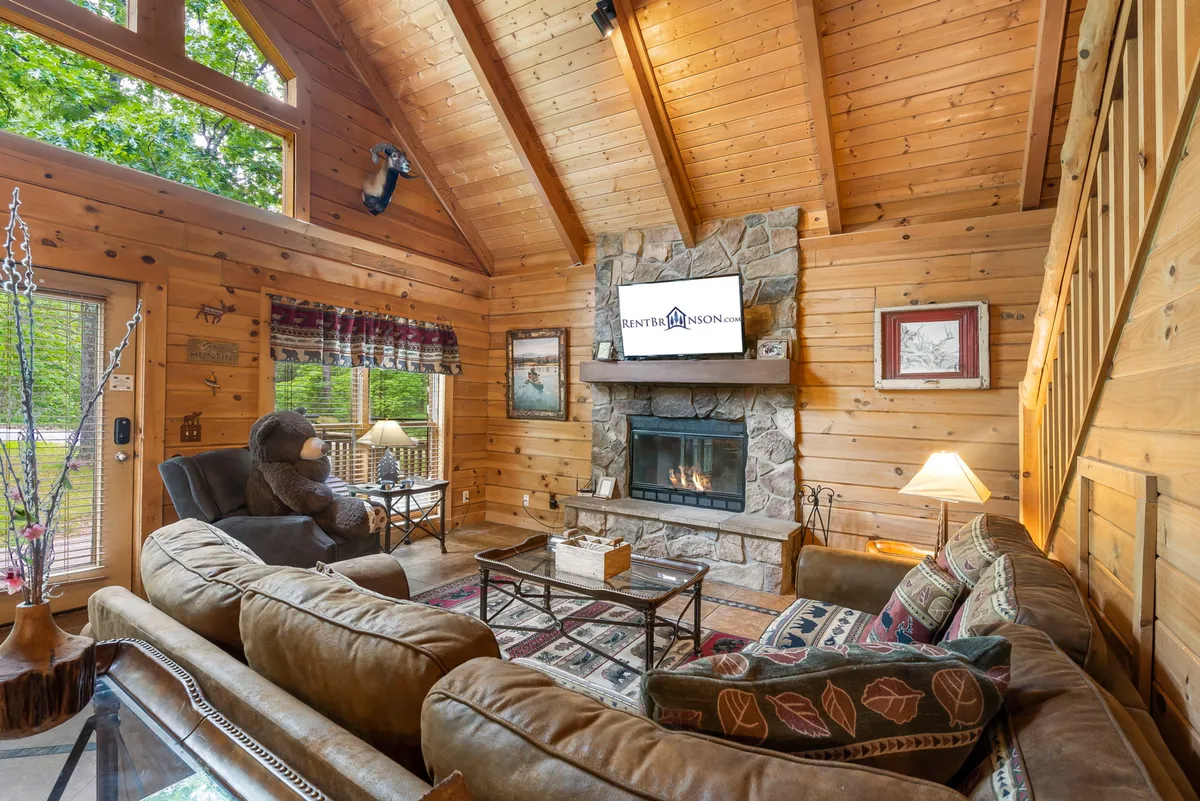 The photo captures a cozy living room centered around a tall stone fireplace. Natural light fills the space through oversized windows, illuminating a comfy sofa, armchair, and a large television mounted nearby. Warm wooden accents and plush seating create an inviting, communal atmosphere.