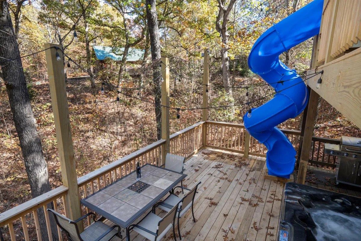 A wide wooden deck with a vibrant blue slide, an outdoor dining table, and Adirondack chairs—all set against a lush natural backdrop. Children are seen playing, and the space is arranged for group dining and family gatherings.