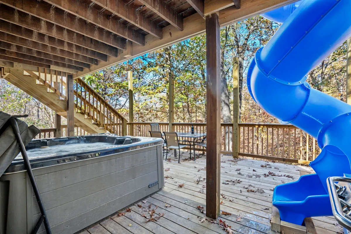 A panoramic exterior view showing a bubbling hot tub, the striking blue slide, a large wooden deck, and the enveloping woods. The scene captures both active play and restful relaxation, with space for gatherings and celebrations.
