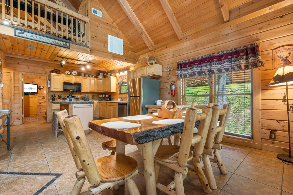 A welcoming dining area features a robust wooden table surrounded by chairs, set next to a kitchen with wooden cabinetry and exposed beams. Large windows bring in forest views and natural light, enhancing the sense of openness and warmth. The setup is ideal for family meals and shared stories.