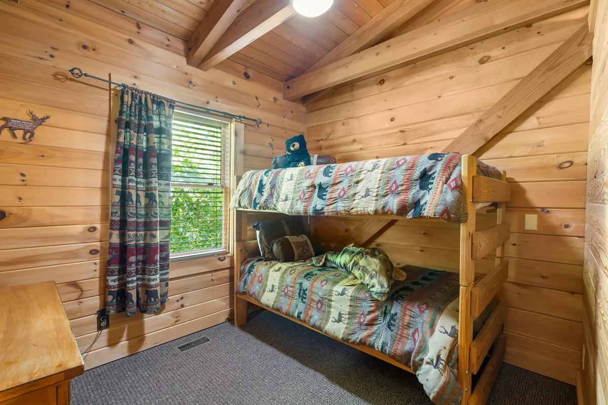 A whimsical bunk room with twin bunks, wooden furnishings, a window with curtains, and a stuffed bear on display. The space is designed for play and relaxation, inviting for kids or teens, and bathed in gentle, natural light.