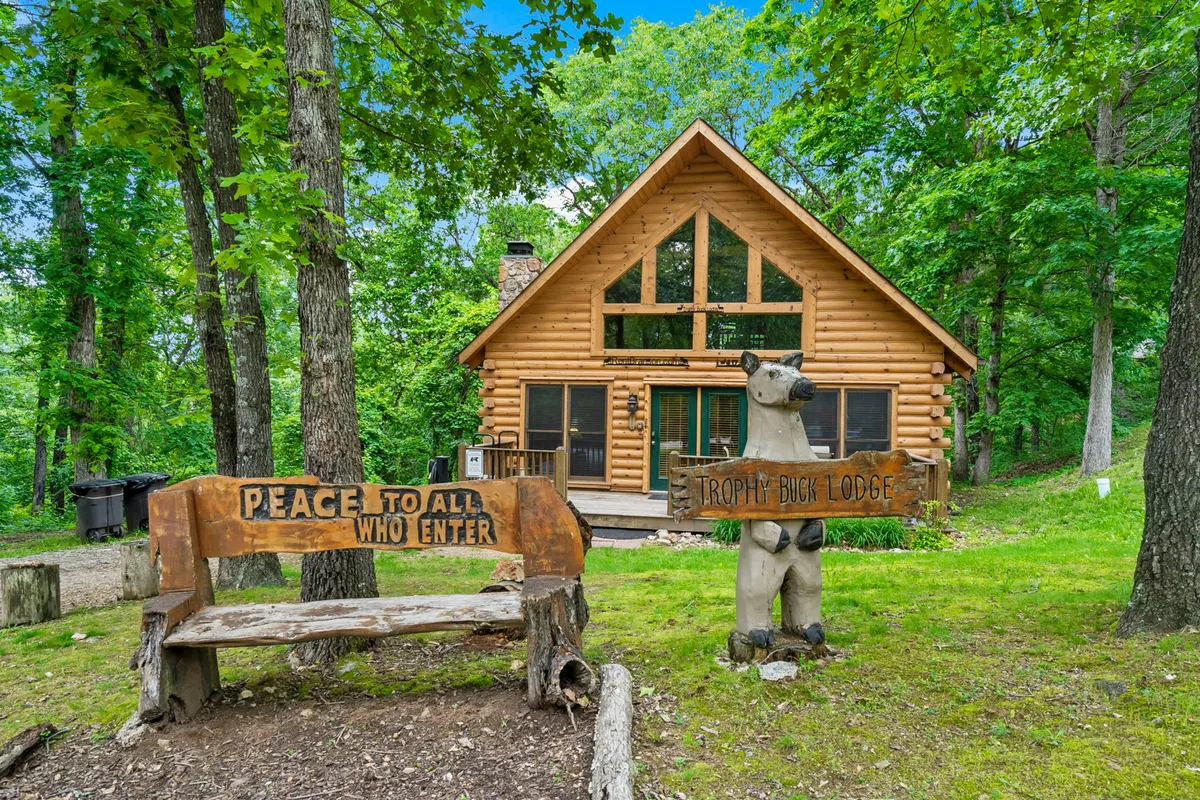 This image shows the log cabin's exterior surrounded by lush greenery, with a welcoming bear statue and a wooden bench with an inscription positioned on the porch. The scene exudes a peaceful, inviting atmosphere, highlighting the cabin's close connection to nature.