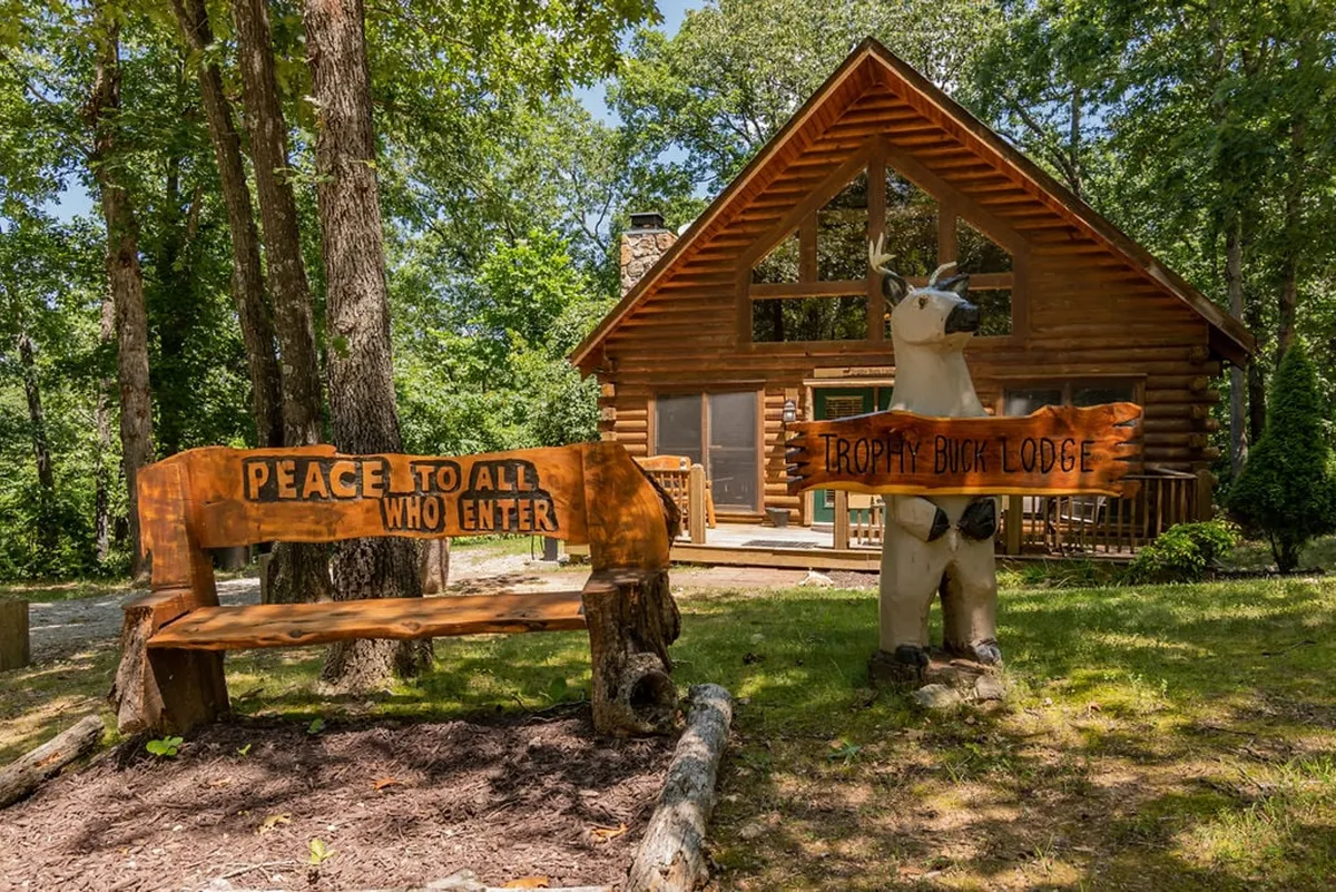 A friendly exterior view of the log cabin, complete with a welcoming wooden bench inscribed with a message and a carved deer statue. The cabin is nestled among trees, suggesting peace and a connection to nature.