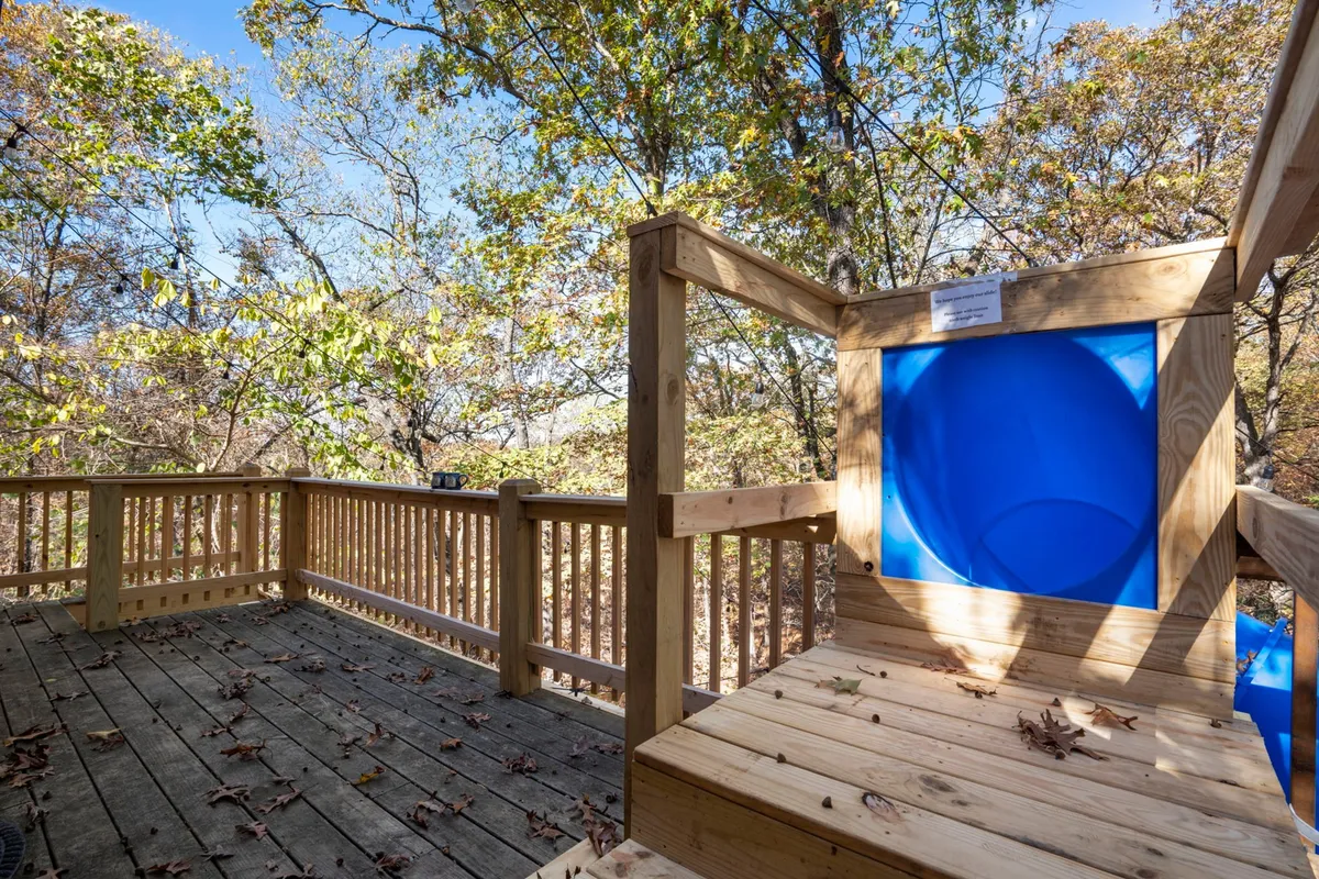 A joyful exterior scene showing a blue slide descending from a wooden deck surrounded by tall trees. Children can be seen playing, while others gather on the deck, suggesting lively family gatherings and opportunities to observe wildlife in the woods.