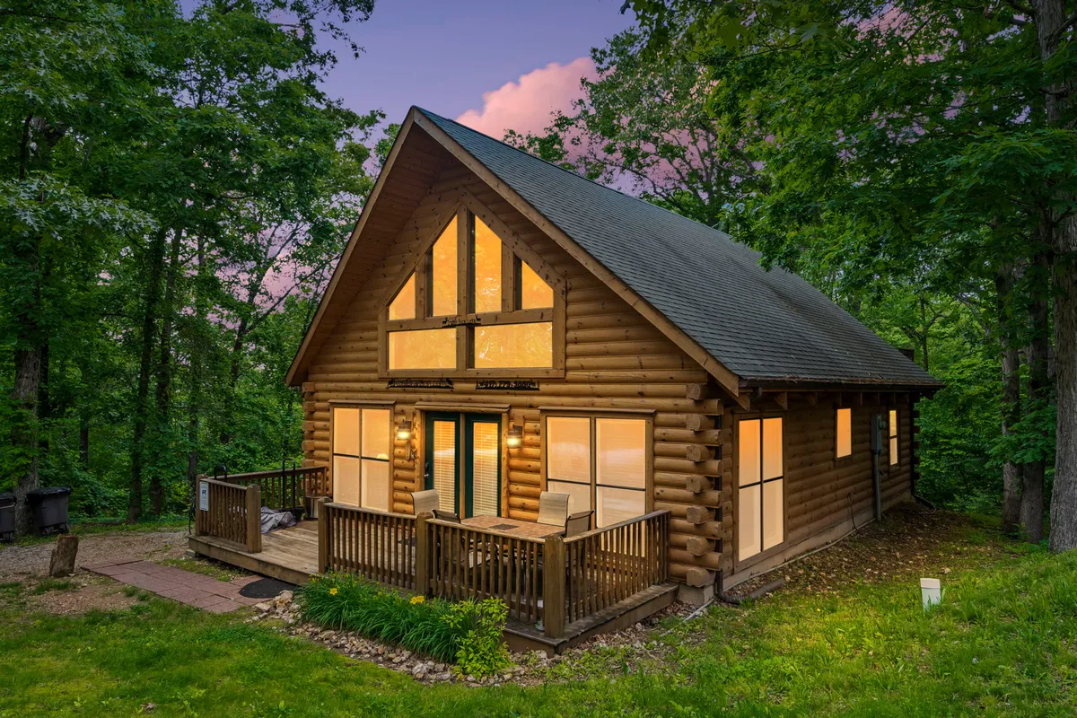 A cozy log cabin with prominent large windows and a spacious wooden deck with seating, set amidst greenery. This inviting exterior highlights the comfortable amenities (deck, seating), forested surroundings, and the seamless blend of indoor comfort and outdoor adventure.