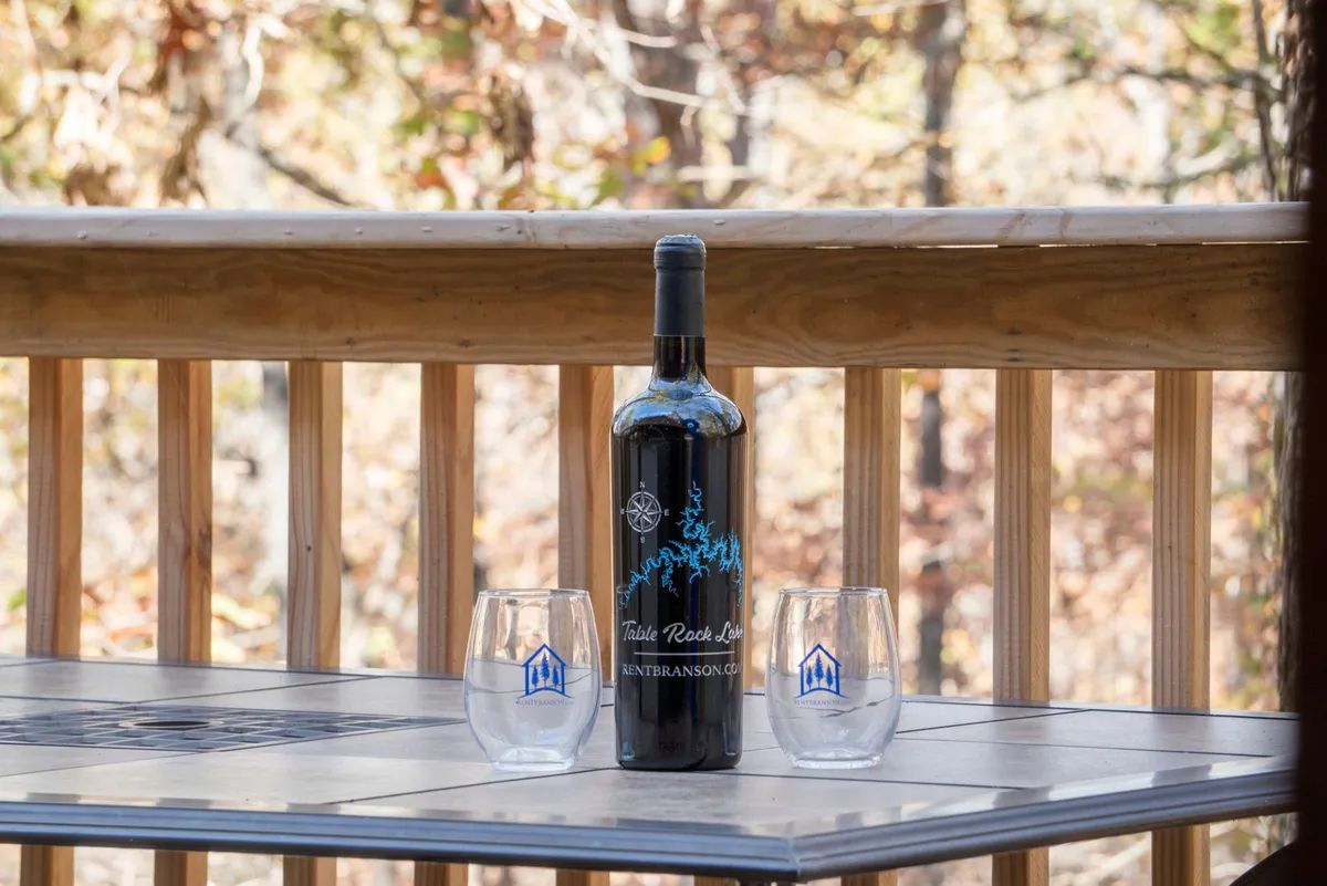 An exterior shot featuring a bottle of wine and two glasses perched on a wooden railing, overlooking a natural setting. The mood is cozy and relaxed, perfect for enjoying evenings outdoors with friends.