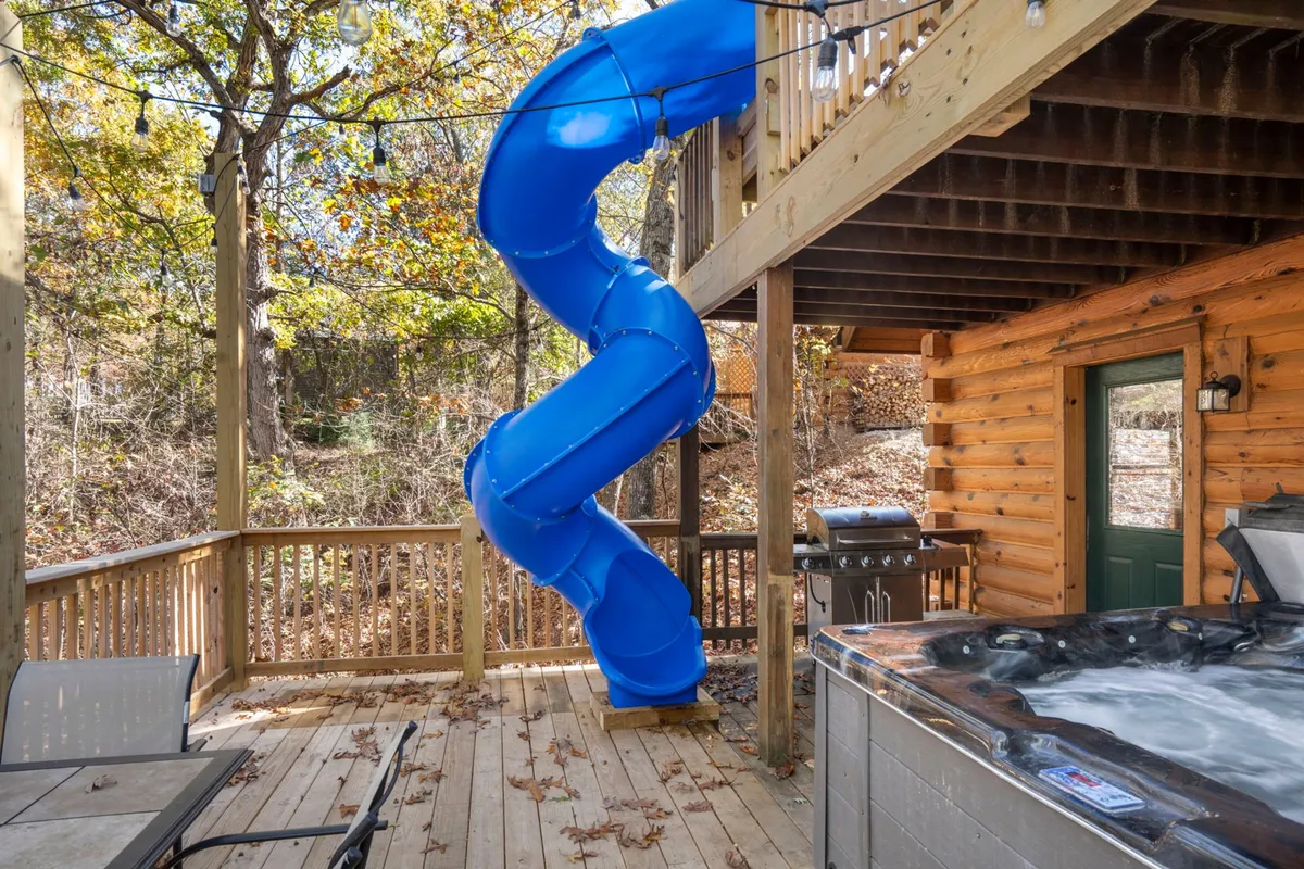 This image captures a spacious wooden deck nestled among trees, featuring a blue water slide, a hot tub, and a grill. The scene includes seating areas and is illuminated by string lights, suggesting a welcoming space for social gatherings, family fun, and relaxing evenings.