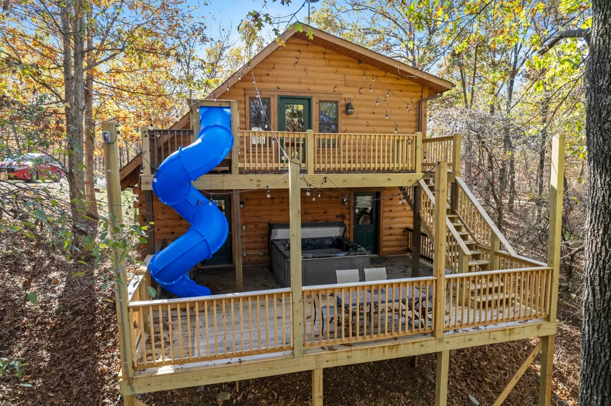 The image shows a charming two-story log cabin nestled among towering Ozark trees, featuring a spacious wooden deck with twinkling fairy lights, a bubbling hot tub, and a vibrant blue slide for playful fun. The tranquil, wooded setting invites relaxation and outdoor gatherings, making it ideal for families or groups seeking a joyful, nature-filled escape.