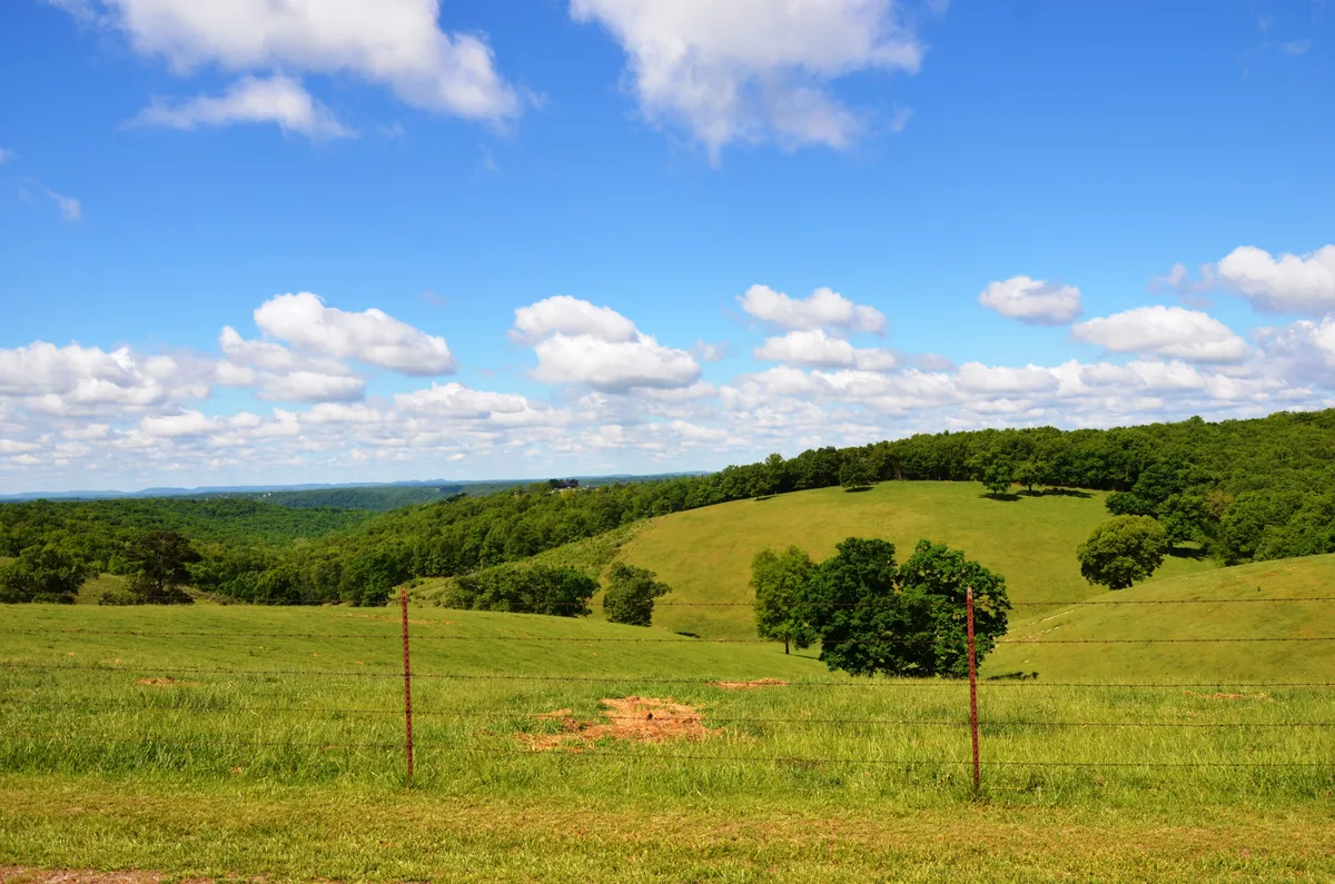 A panoramic view of rolling hills, green trees, and a blue sky dotted with clouds, framed by a simple fence line. The scene conjures the vast, scenic Ozark landscape, evoking hiking adventures and moments of outdoor reflection.