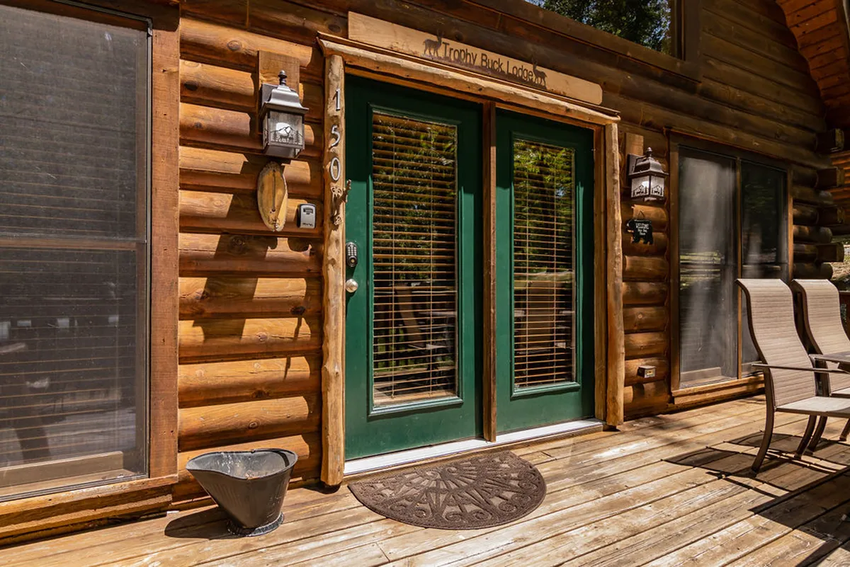 This exterior shot highlights the log cabin's double green doors, flanked by decorative lanterns, with a cozy welcome mat at the entrance. The scene feels inviting and homely, emphasizing the warm welcome guests receive.