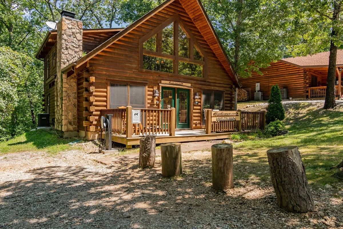 A log cabin with a spacious front deck set amidst vibrant greenery, featuring wooden posts and a stone chimney. The deck invites relaxation and provides a transition between indoor comfort and the outdoor experience, ideal for unwinding after a day of hiking with your pet.