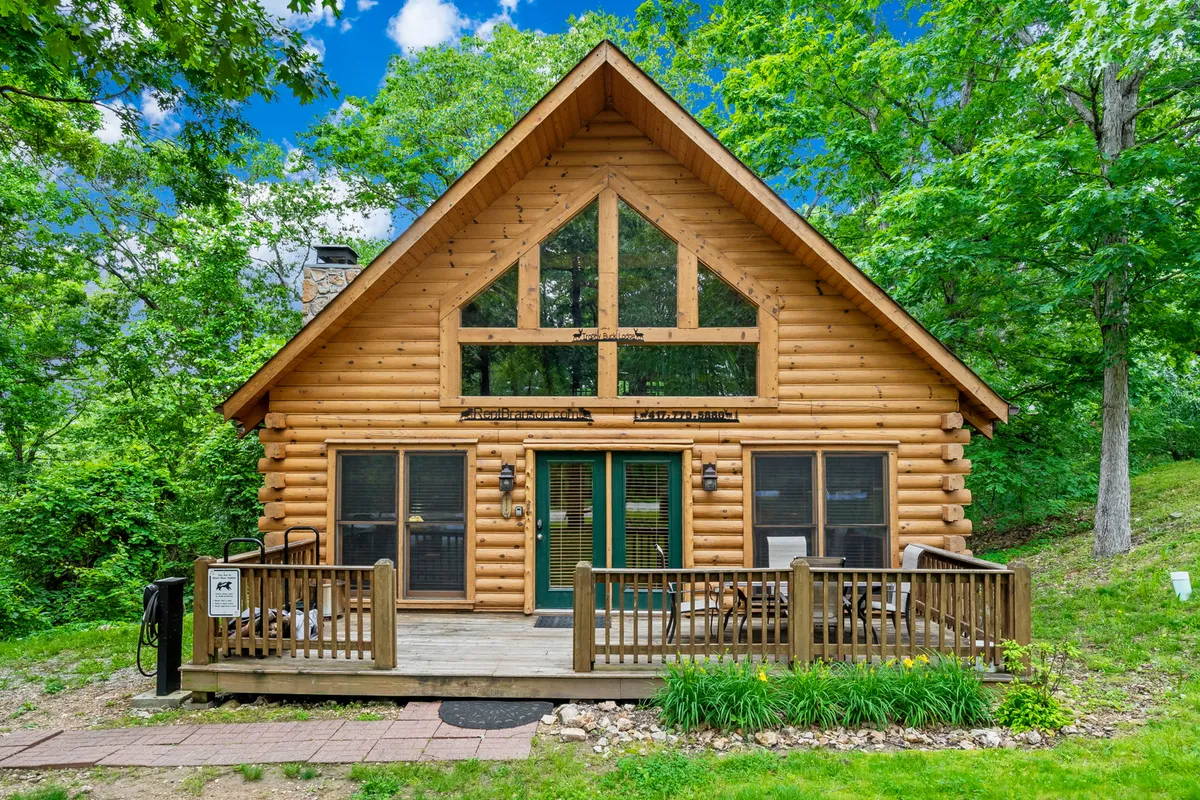 This exterior image shows the log cabin with large front windows, a spacious wooden deck, and lush greenery surrounding the home. The setting suggests a welcoming atmosphere ideal for group gatherings, relaxing outdoors, and enjoying the natural environment.